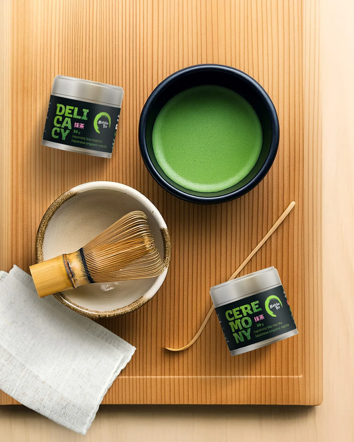 Matcha po šedesátce: co ukázaly studie a jak nápoj správně připravit 1 Ceremoniální sada na přípravu matchy: dózy Delicacy a Ceremony, tmavá miska chawan s napěněným nápojem, bambusová metlička chasen a lžička chashaku na dřevěném prkénku
