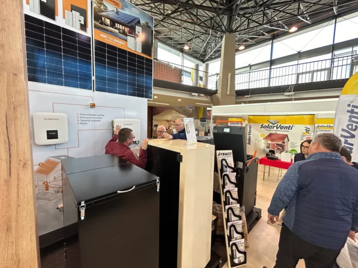 Expozice na veletrhu úspor: fotovoltaické panely, tepelné čerpadlo Dražice a zásobník na pelety, u pultu se návštěvníci baví s vystavovatelem