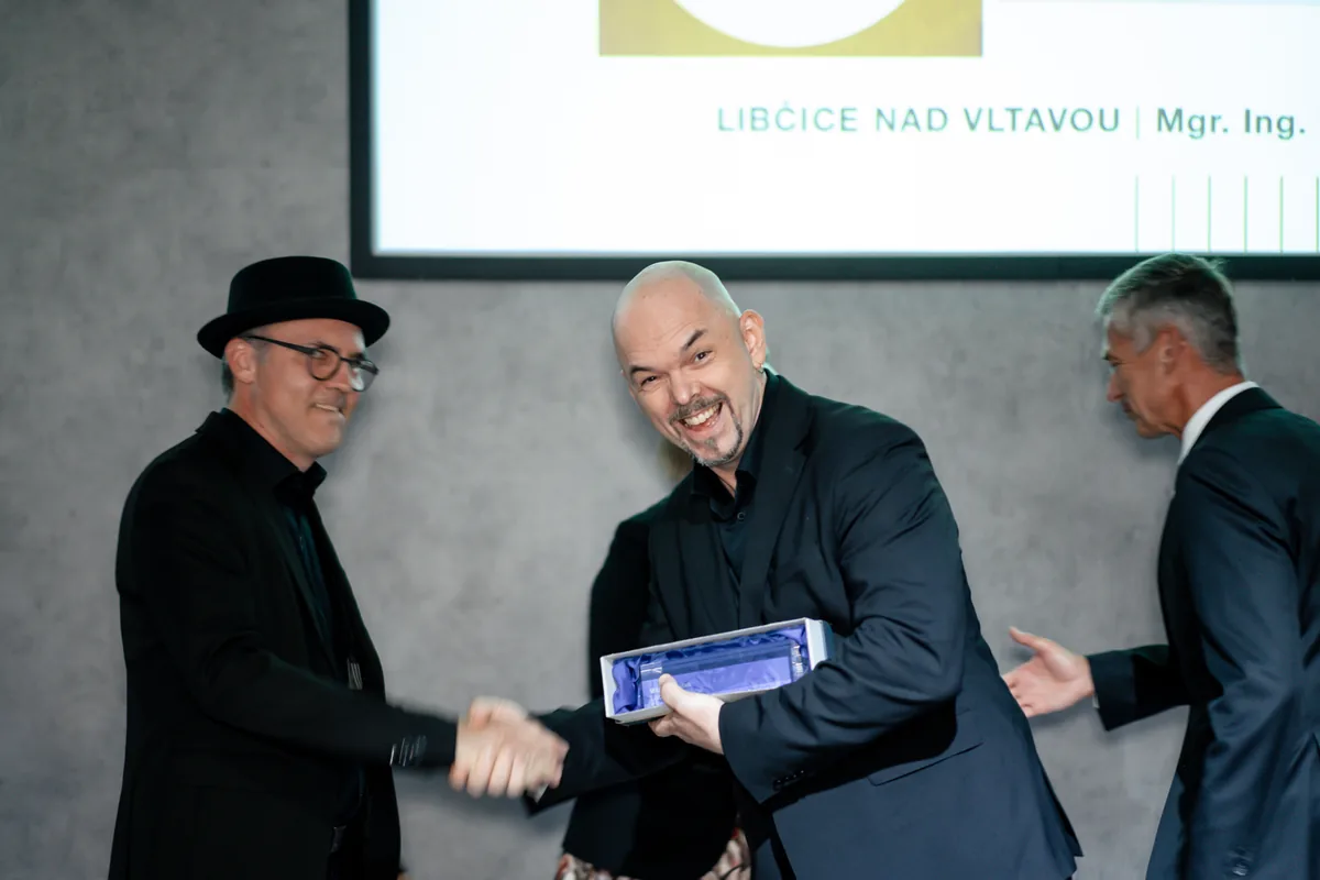 Architekt roku 2026 získali Ehl a Koumar, Architekt obci Libčice 2 Milan Kopeček přebírá cenu Architekt obci 2026 v dárkové krabici a podává si ruku s předávajícím, v pozadí plátno s nápisem Libčice nad Vltavou